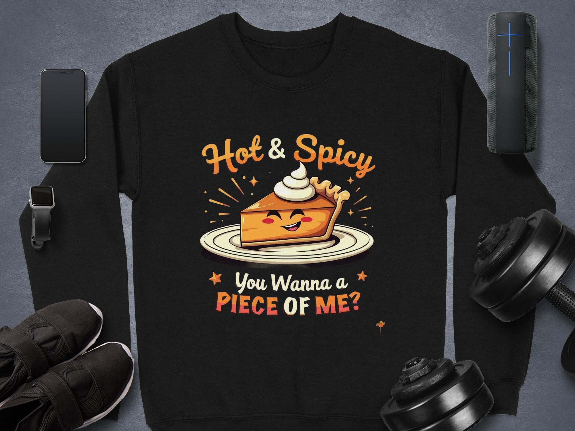 A Garment Graphics black sweatshirt features a playful pie character with sunglasses and whipped cream, reading Hot & Spicy. You Wanna a Piece Of Me? Its unisex and surrounded by a smartphone, smartwatch, sneakers, speaker, and dumbbells.