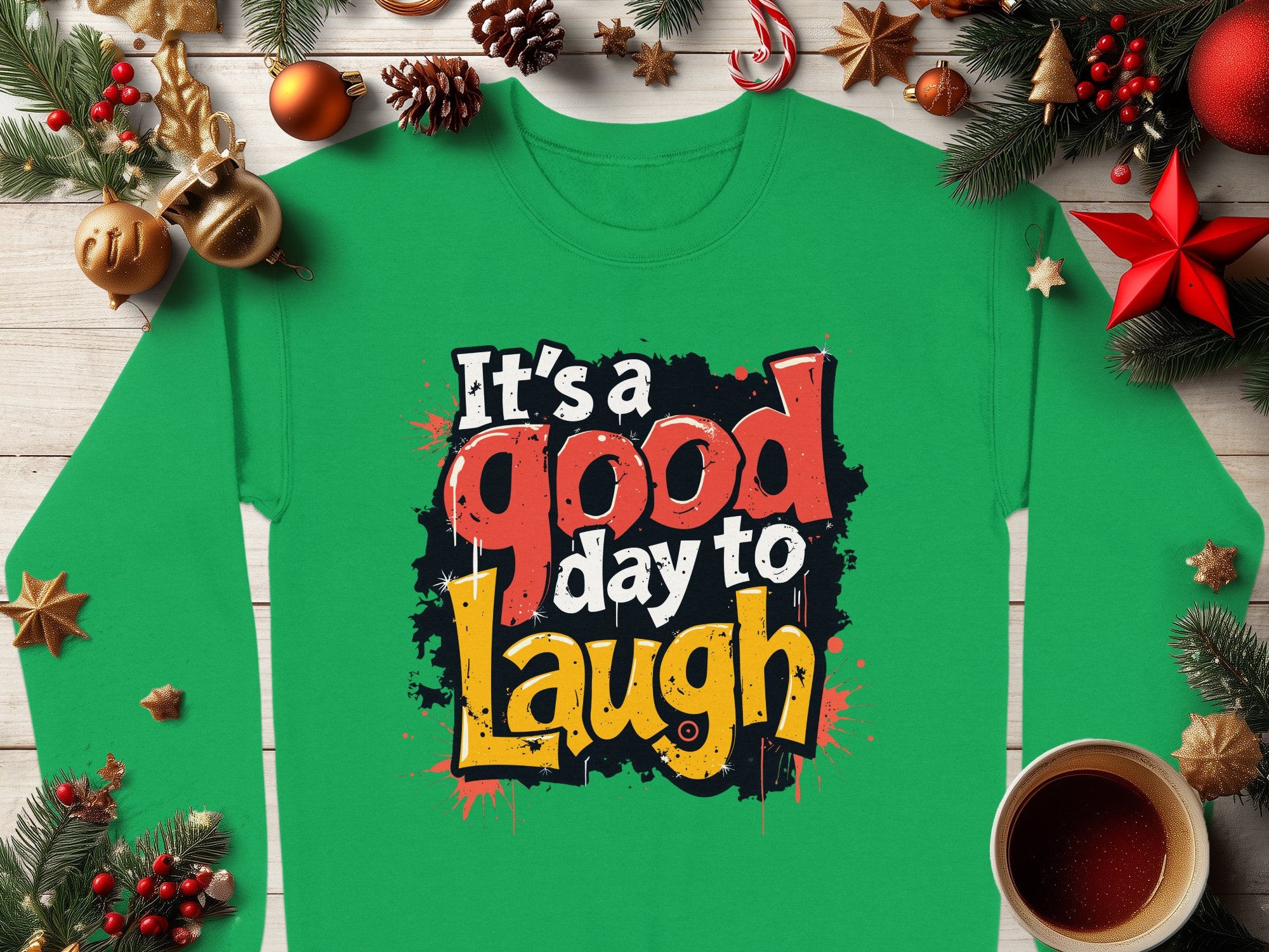 A unisex Garment Graphics green sweatshirt features Its a Good Day to Laugh in colorful, bold letters, surrounded by Christmas decorations, pine branches, ornaments, and a cup of hot chocolate on a wooden surface.