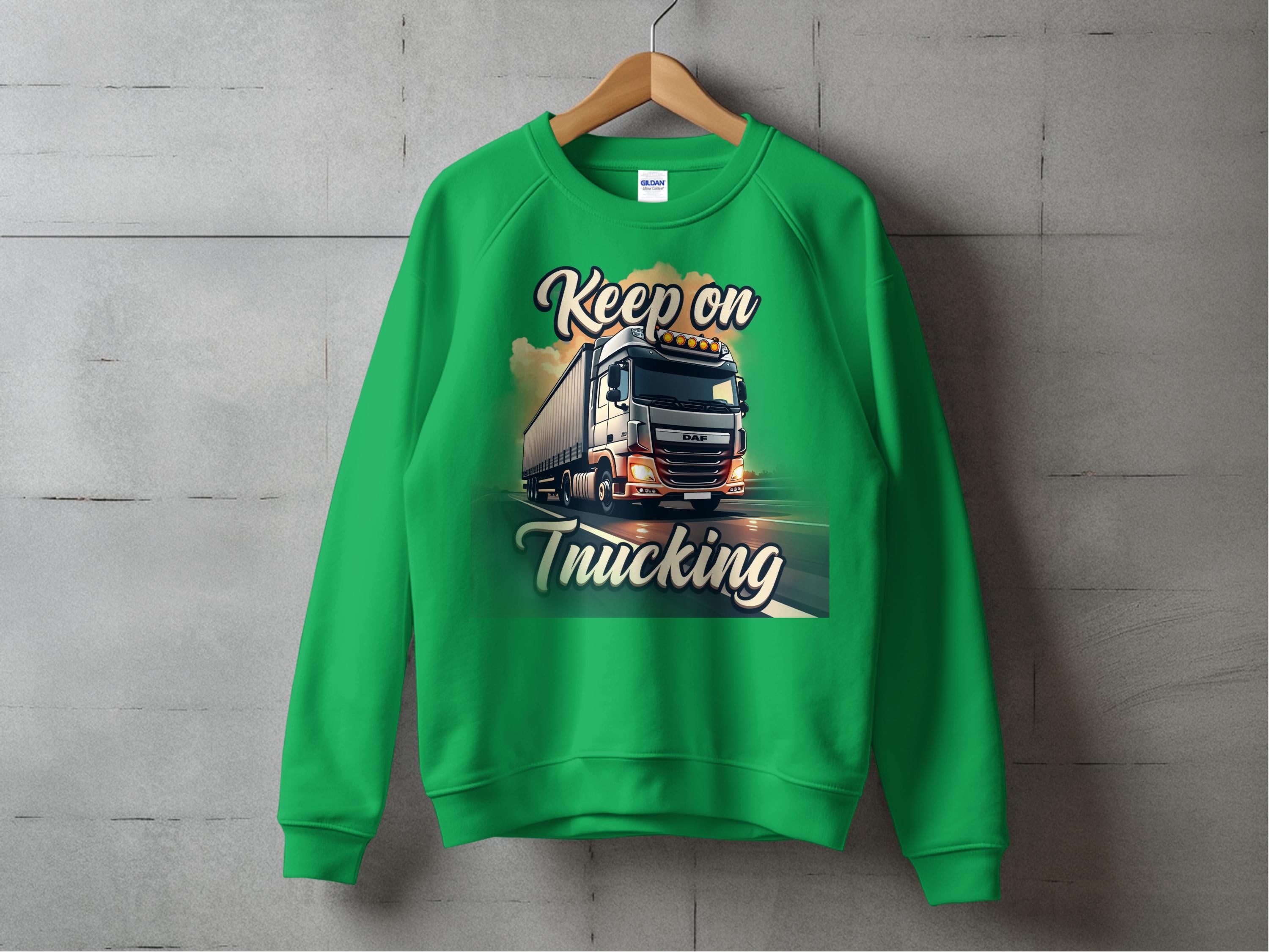 Garment Graphics inspirational green sweatshirt features a truck and Keep on Trucking design. With light and clouds framing the forward-moving truck, it embodies the open road spirit against a gray wall.