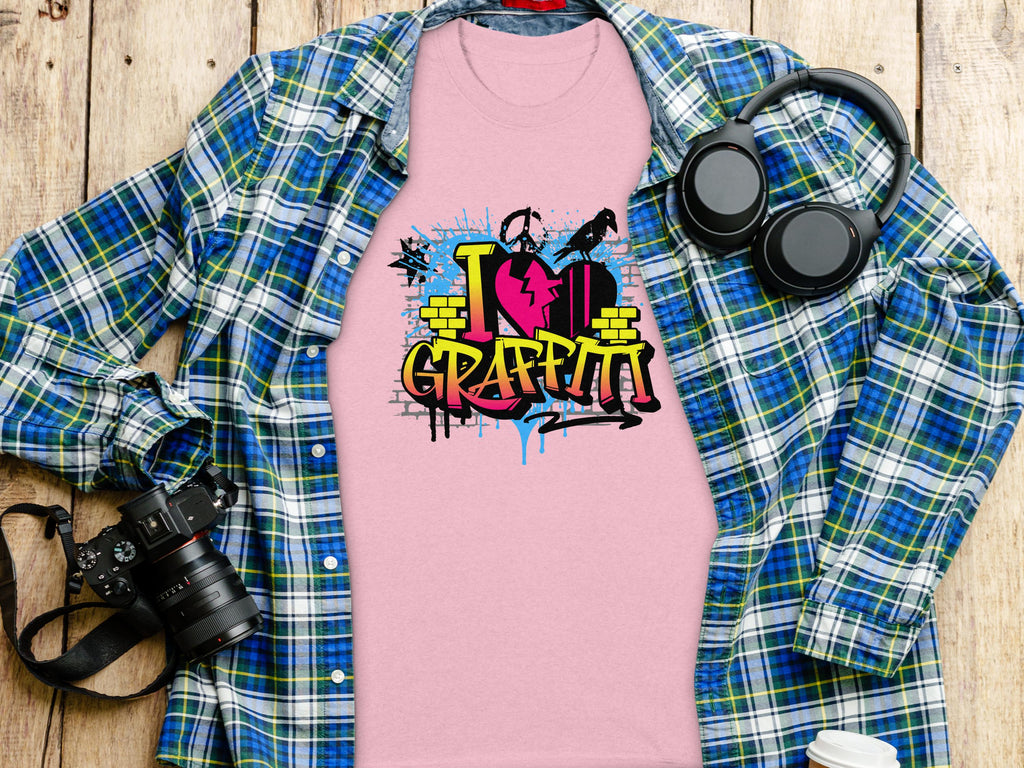 A pink Graffiti Love T-Shirt by Garment Graphics, featuring a vibrant I Love Graffiti street art design, is on a wooden surface partially covered by a blue and green plaid shirt. Nearby are a camera, headphones, and a white cup with a black lid.