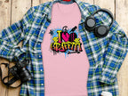 A pink Graffiti Love T-Shirt by Garment Graphics, featuring a vibrant I Love Graffiti street art design, is on a wooden surface partially covered by a blue and green plaid shirt. Nearby are a camera, headphones, and a white cup with a black lid.