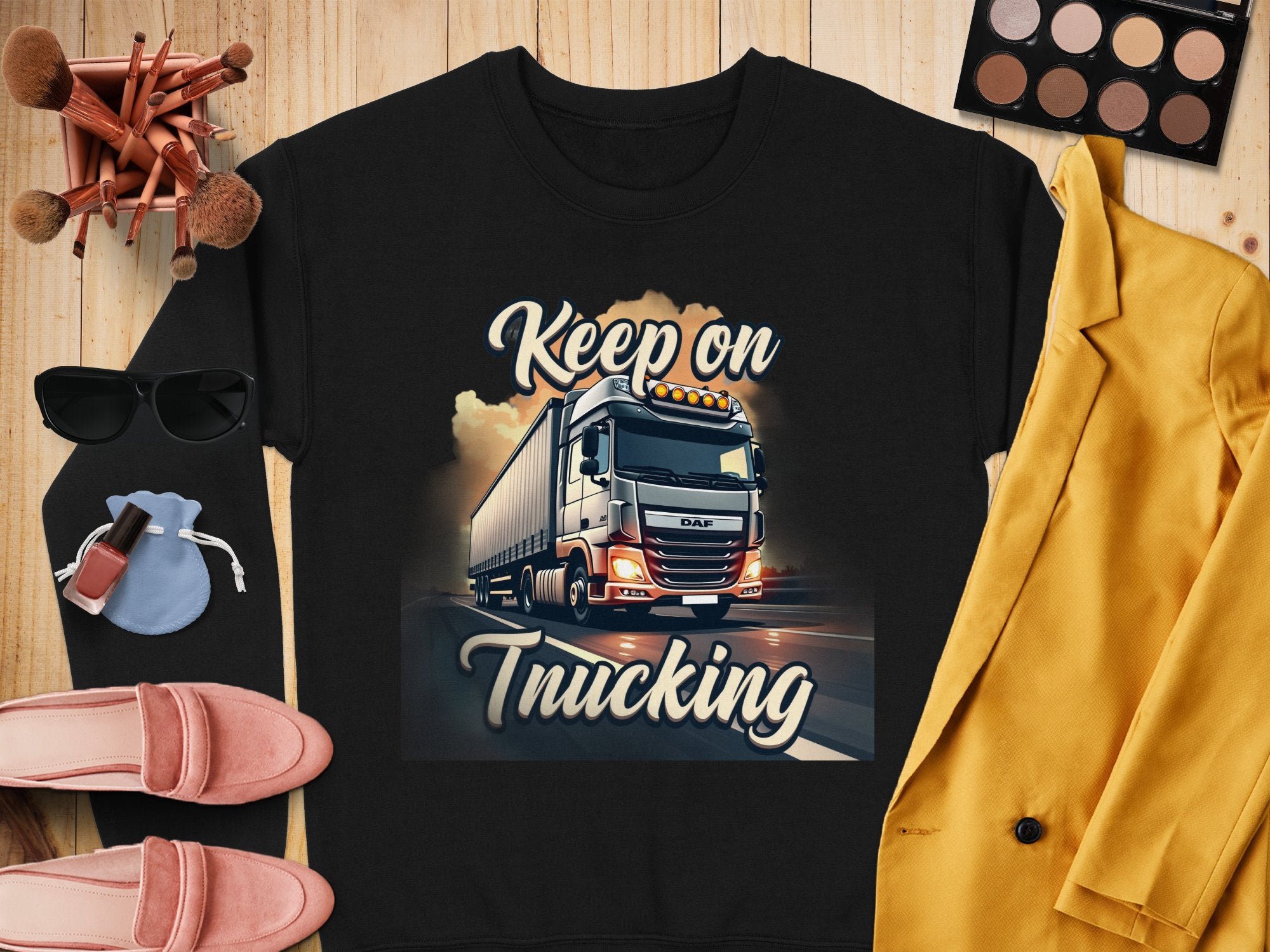 Garment Graphics graphic sweatshirt showcases a Keep on Trucking text and vibrant semi-truck design. Styled with makeup, brushes, sunglasses, pink shoes, a blue pouch, and a yellow jacket on a wooden surface for a relaxed vibe.