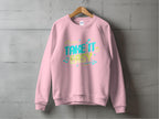 A pink unisex sweatshirt from Garment Graphics hangs on a wooden hanger against a gray concrete wall, featuring colorful Take It Easy text with decorative arrows and musical notes.