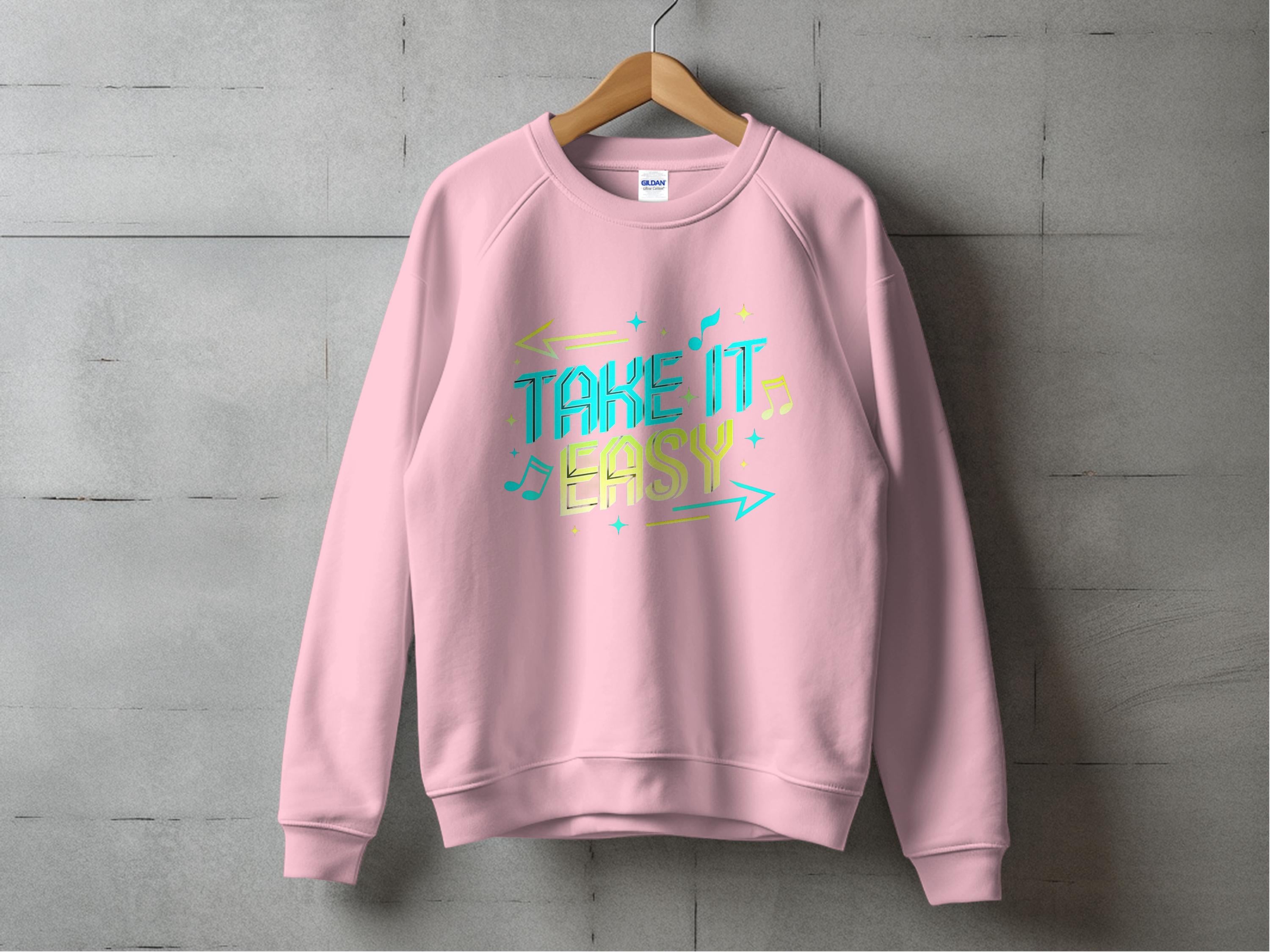 A pink unisex sweatshirt from Garment Graphics hangs on a wooden hanger against a gray concrete wall, featuring colorful Take It Easy text with decorative arrows and musical notes.