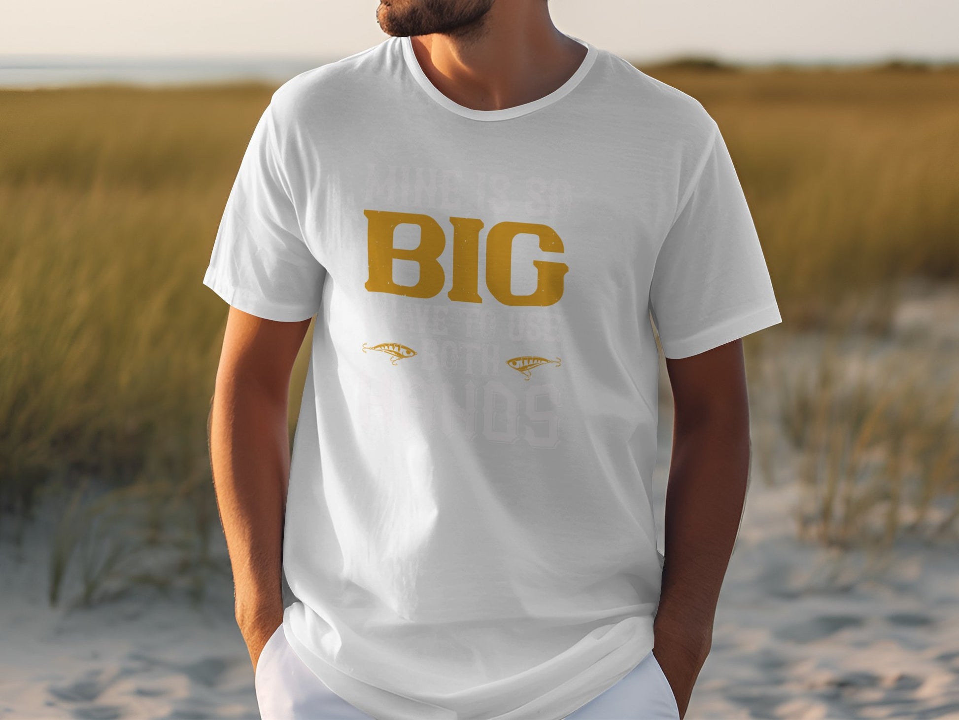 A person wearing a Garment Graphics white graphic tee featuring BIG in large gold letters stands against a sandy beach backdrop with blurred grass. Hands casually in their pockets, they embrace the laid-back vibe of classic fit apparel.