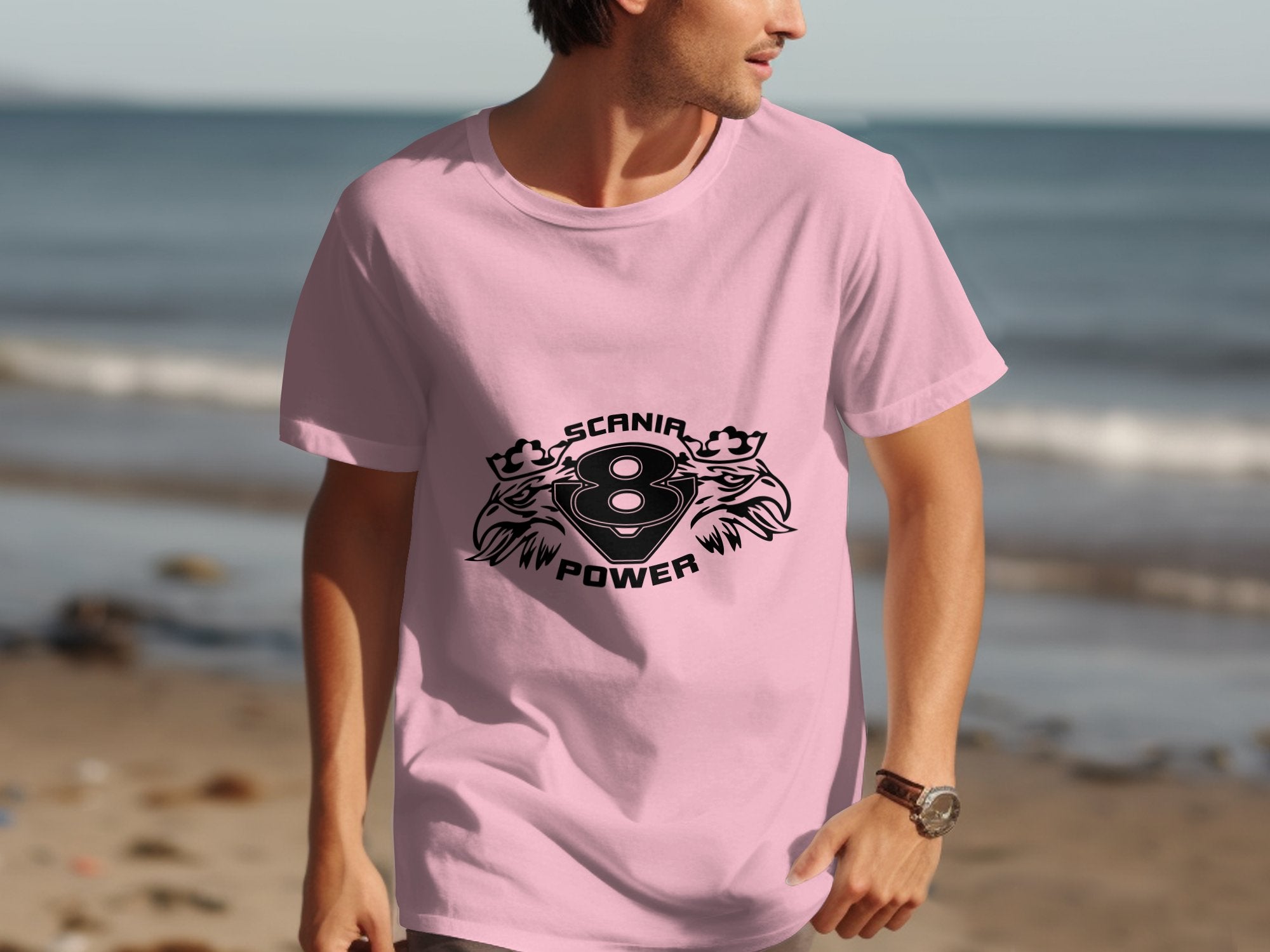 A man wearing a vibrant pink Scania Power Eagle graphic tee from Garment Graphics strolls along the beach, where the serene ocean beautifully contrasts with the sandy shore.