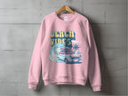 A Garment Graphics pink unisex sweatshirt hangs on a concrete wall, boasting a retro roadtrip design with a classic car, surfboard, palm trees, ocean waves, and bold BEACH VIBES text.