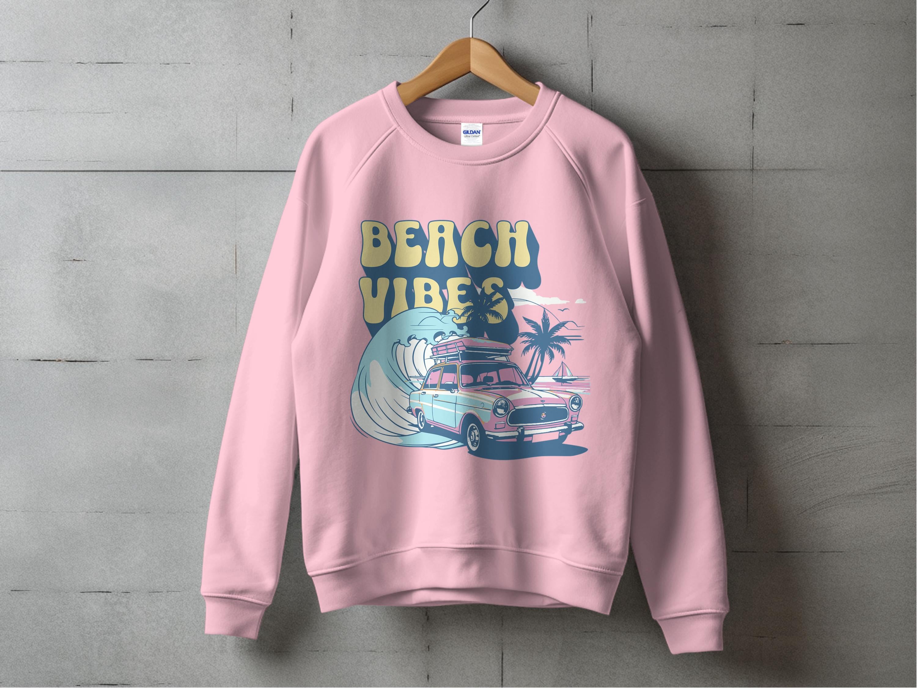 A Garment Graphics pink unisex sweatshirt hangs on a concrete wall, boasting a retro roadtrip design with a classic car, surfboard, palm trees, ocean waves, and bold BEACH VIBES text.