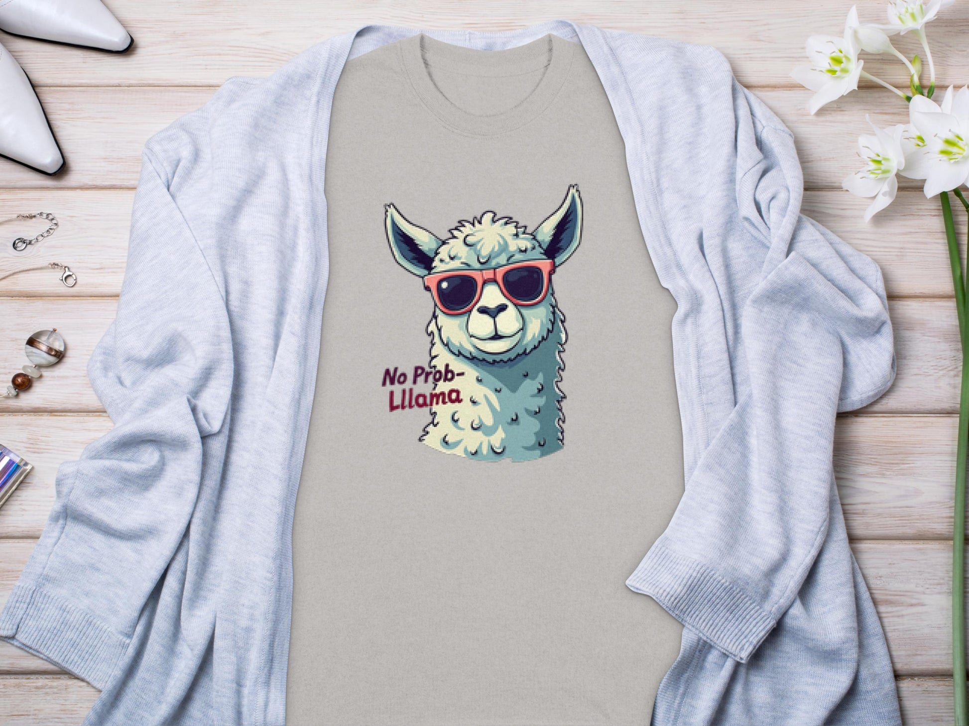 A Garment Graphics beige Llama T-shirt, ideal for families, showcases a whimsical llama with red sunglasses and No Prob-Llama text. Its styled flat on a light gray cardigan with jewelry and white flowers.