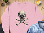 A bold, pink sweatshirt by Garment Graphics featuring a large skull and crossbones graphic is laid on a wooden table. Nearby are a detailed latte, small potted plant, denim jacket, and dark textured fabric.