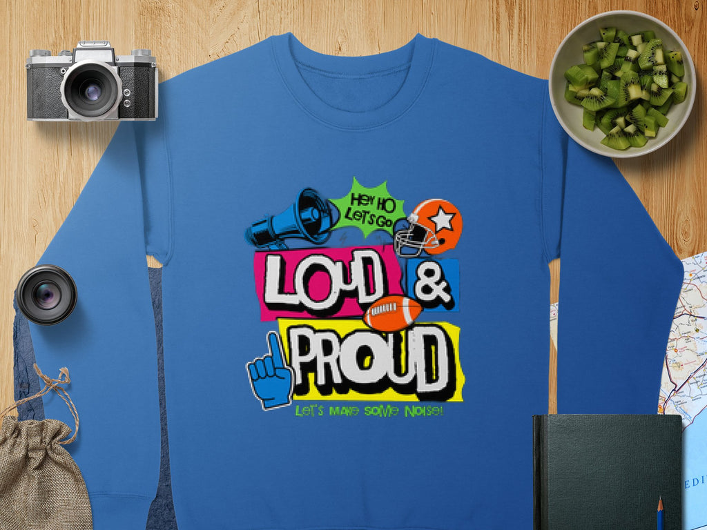 On a wooden table lies a unique unisex blue sweatshirt from Garment Graphics, featuring a vibrant design of a megaphone, foam finger, and football helmet with the text Hey Ho Lets Go Loud & Proud Lets Make Some Noise! Nearby are a camera, lens, map, book, and bowl of kiwi.