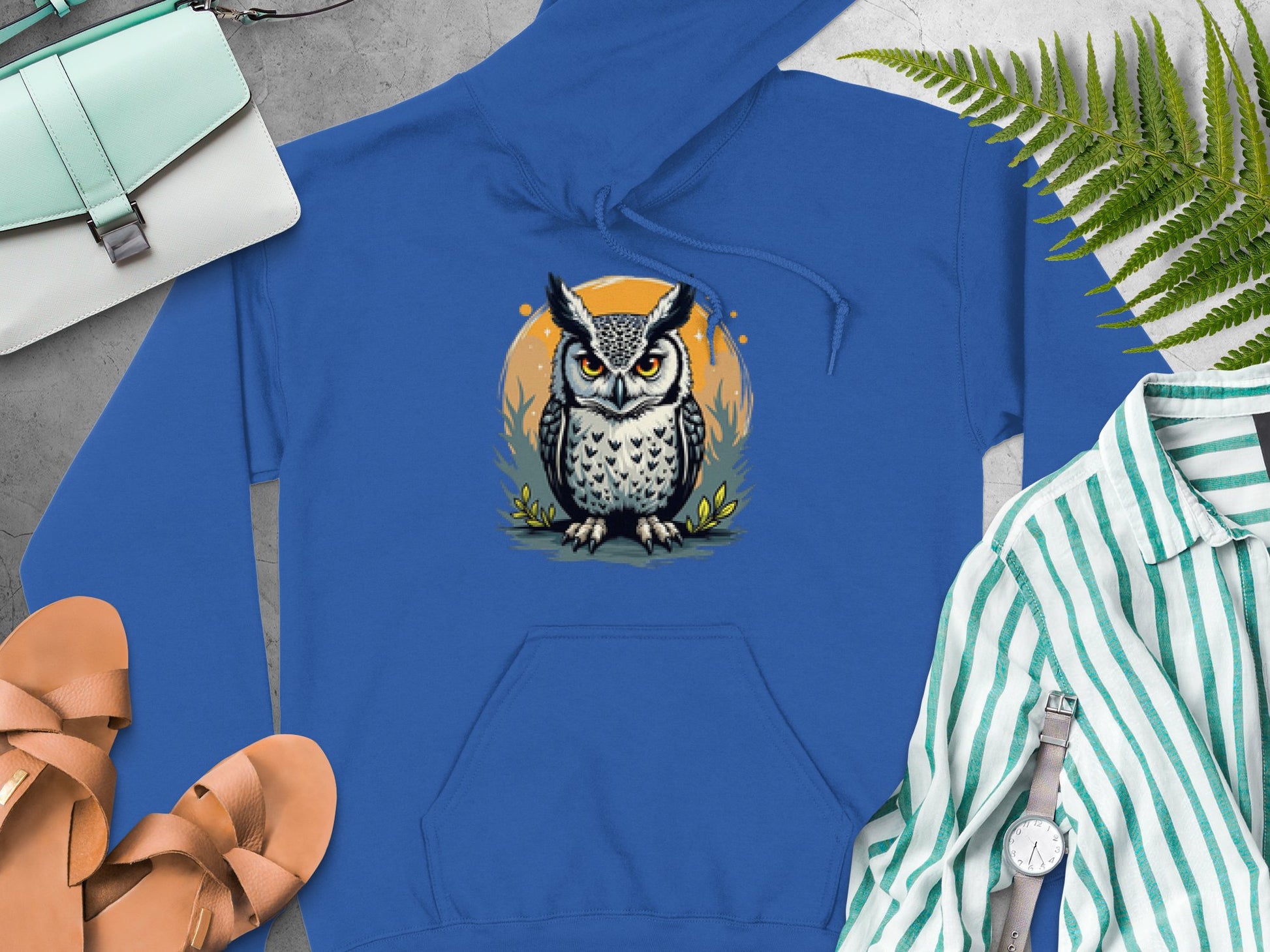 The Garment Graphics Fierce Owl Illustration Hoodie, made from medium-heavy fabric, showcases a cartoon owl on grass with an orange background. It pairs perfectly with tan sandals and a white purse, set against green ferns on concrete for a distinctive style boost.