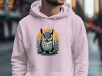 A person in a Garment Graphics classic fit hoodie is outdoors in an urban setting. The medium-heavy fabric features an owl with vivid eyes on a yellow-orange circle with leaves. Buildings and blurred pedestrians are visible in the background.