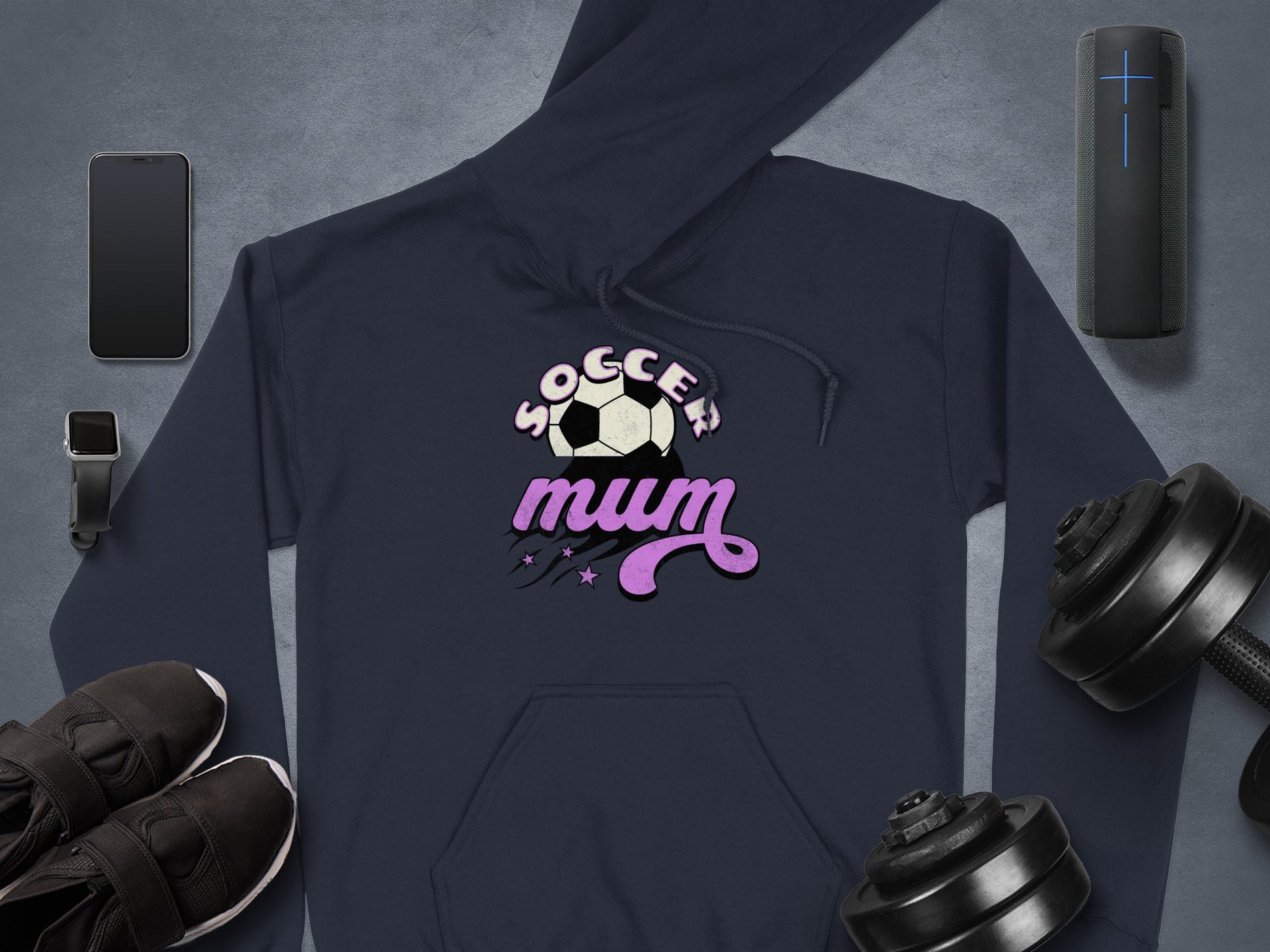 A Garment Graphics hoodie, featuring Soccer Mum text and a soccer ball graphic, is displayed on a gray surface. This cozy classic-fit piece is surrounded by essentials like a smartphone, smartwatch, earbuds, portable speaker, sneakers, and dumbbells.