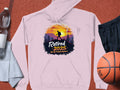 A Garment Graphics pink hiking hoodie with a sunset mountain and hiker design features the text Retired 2025: Not My Problem Anymore. It lies on a vibrant red background, surrounded by a basketball, water bottle, towel, and black shorts.