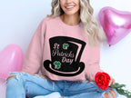 A woman with long wavy hair celebrates in a Garment Graphics pink sweatshirt, featuring a black top hat, shamrocks, and St Patricks Day text. Holding a red rose, she sits against a white background with pink heart balloons, wearing ripped jeans for added casual charm.