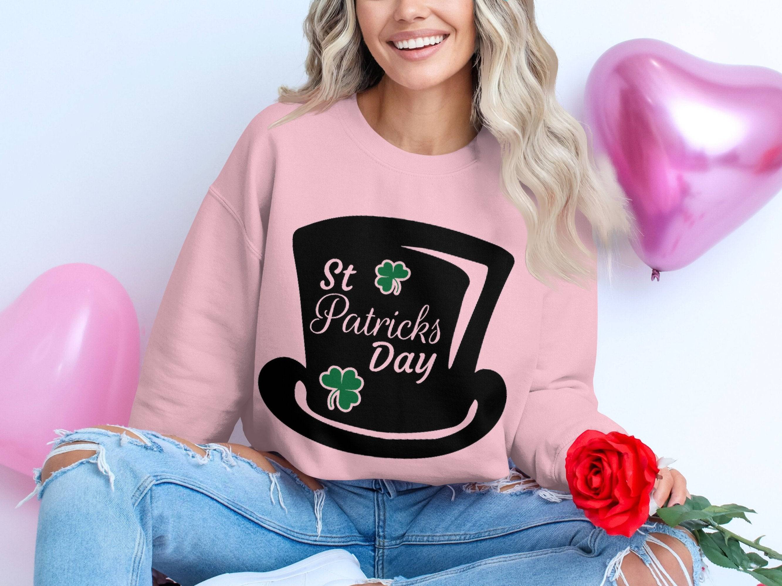 A woman with long wavy hair celebrates in a Garment Graphics pink sweatshirt, featuring a black top hat, shamrocks, and St Patricks Day text. Holding a red rose, she sits against a white background with pink heart balloons, wearing ripped jeans for added casual charm.