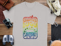 A light gray t-shirt by Garment Graphics lies on a wooden surface, featuring a Woodstock 1969 retro design with a red bird and van on a rainbow background. Perfect for music lovers, its surrounded by a camera, binoculars, leather bag, and gloves.