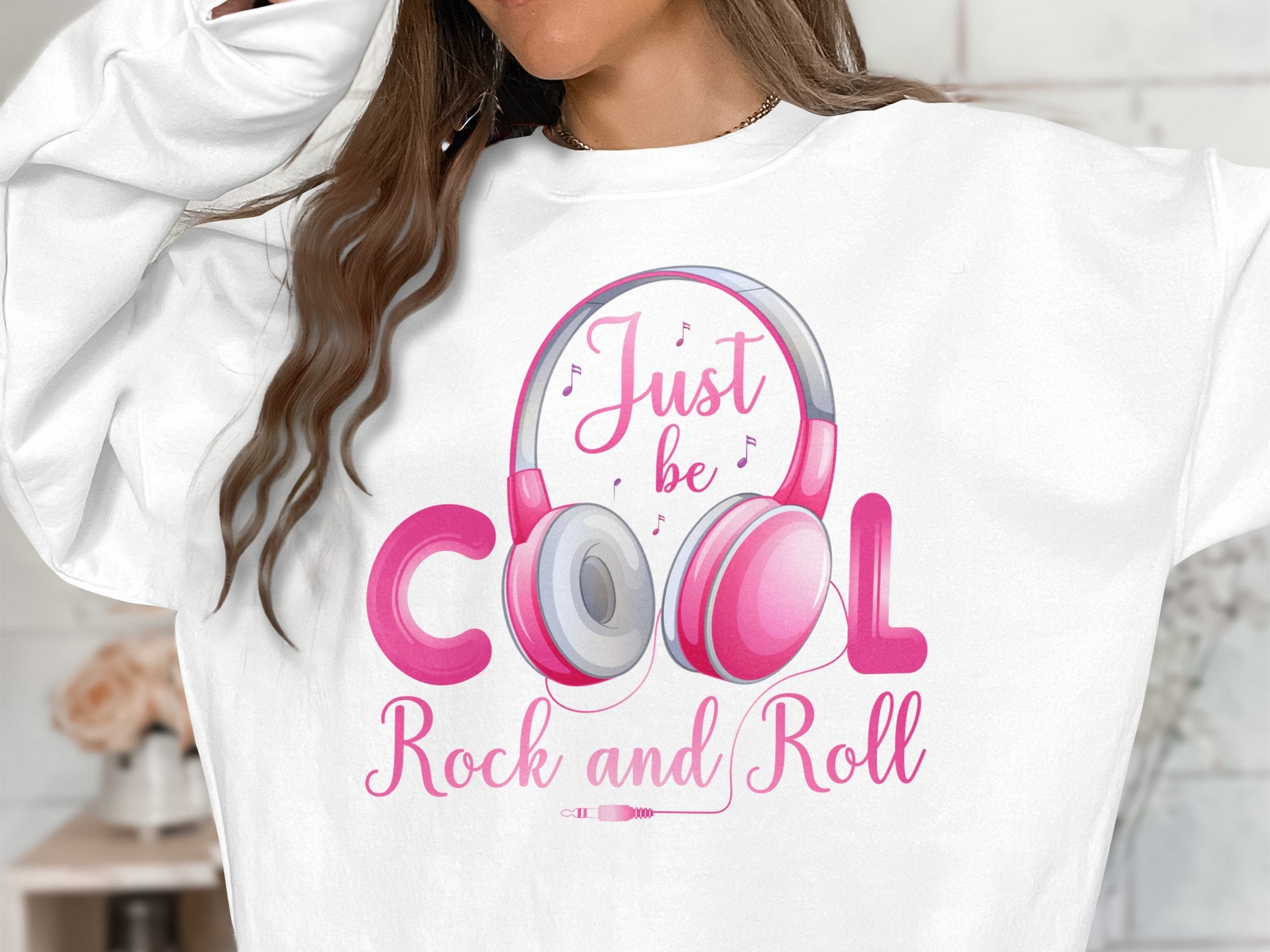 A person sports a fashionable white Garment Graphics sweatshirt, embellished with pink Rock and Roll headphones, musical notes, and the phrase Just be Cool Rock and Roll. Their long, wavy brown hair adds flair to their music-inspired style against a blurred backdrop.