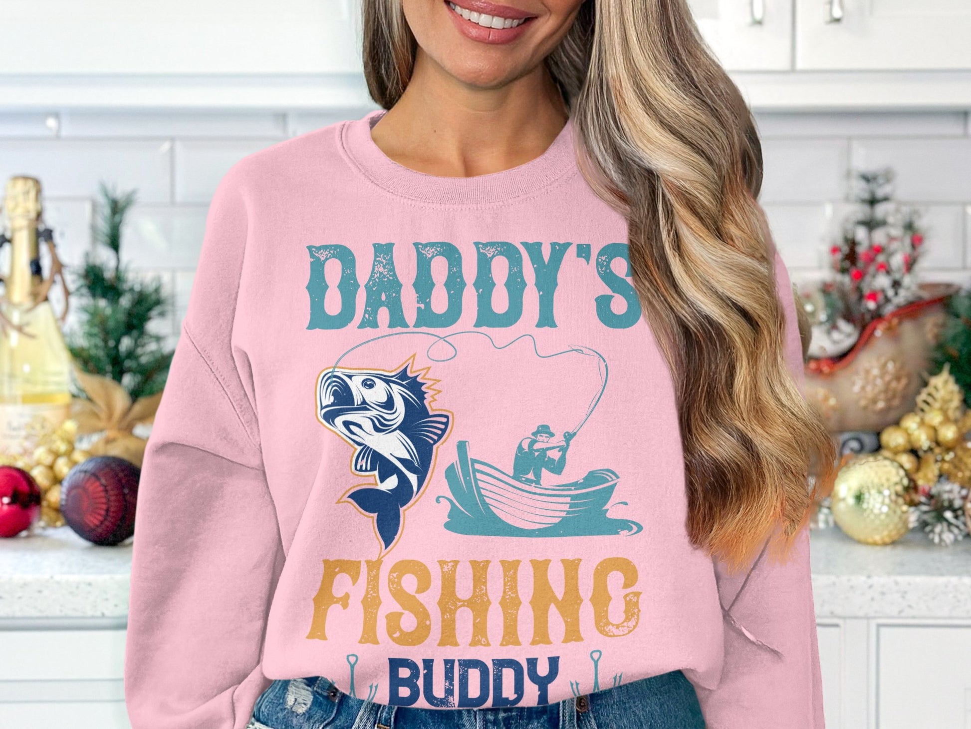 A woman stands in a festive kitchen wearing Garment Graphics pink Daddys Fishing Buddy sweatshirt, featuring a fish and person in a boat illustration. Holiday decor enhances the atmosphere for fishing fans.