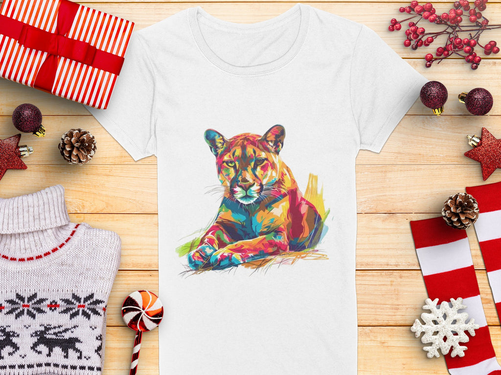 A white Garment Graphics T-shirt with a colorful puma design sits on a wooden surface, surrounded by wrapped gifts, pinecones, a candy cane, a folded sweater, and festive decorations like snowflakes and stars.