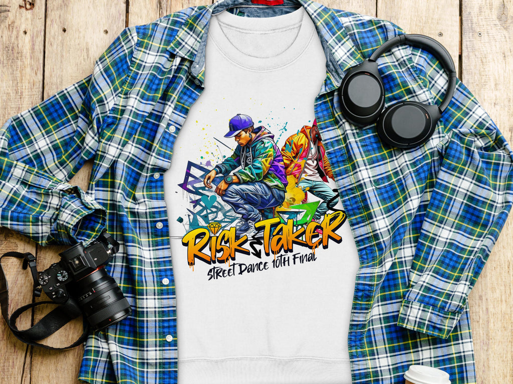 A Garment Graphics white t-shirt featuring Risk Taker Street Dance 10th Final and colorful urban-style dancer graphics is laid on a blue plaid shirt. Nearby are black headphones, a camera, and a white cup on wood. Available in unisex sizing for street dance fans.