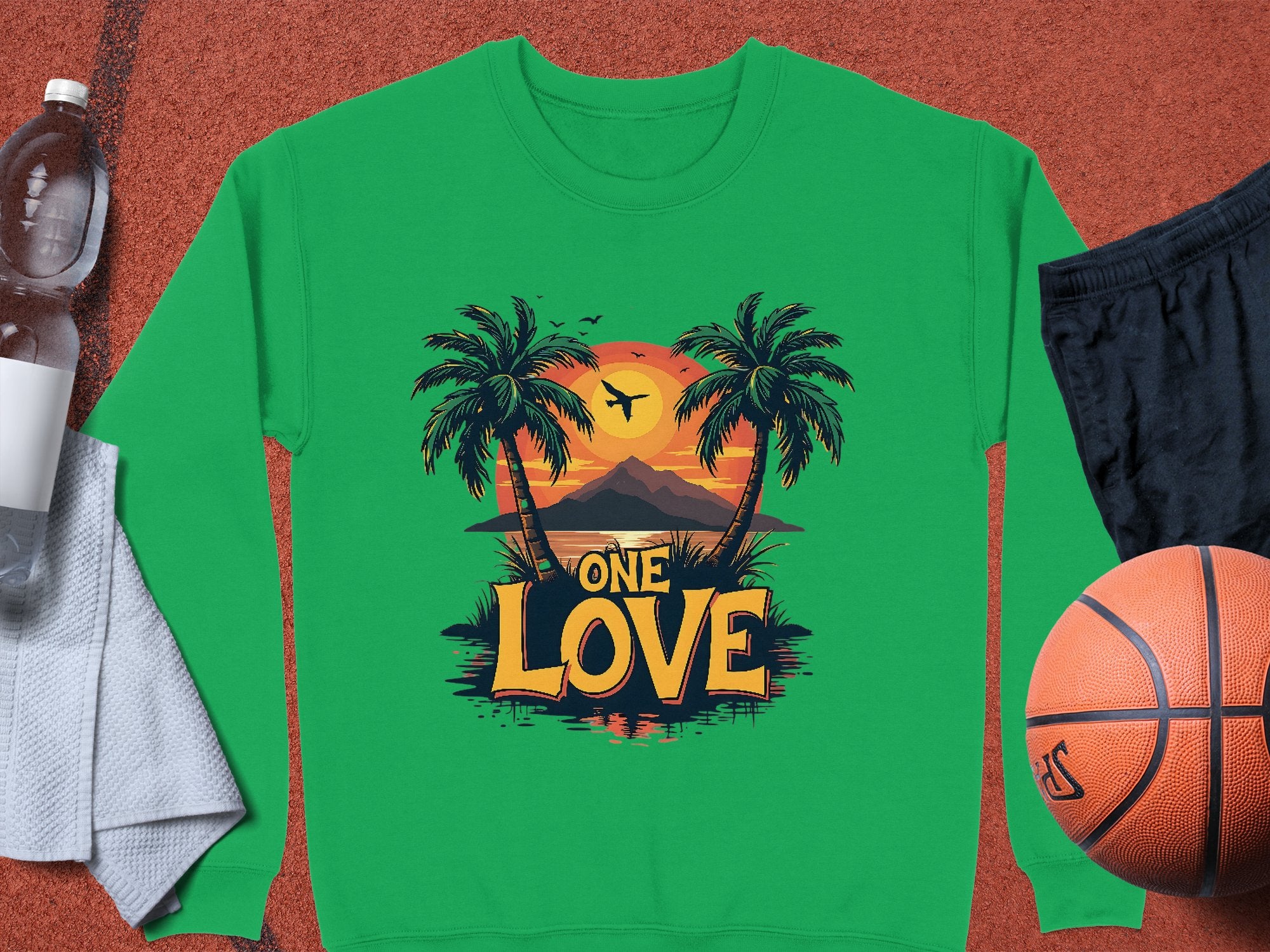 A green sweatshirt from Garment Graphics One Love Sweatshirts collection displays a vibrant sunset, palm trees, mountain, and plane with One Love text. Unisex sizing makes it a perfect fit next to a water bottle, towel, black shorts, and basketball.
