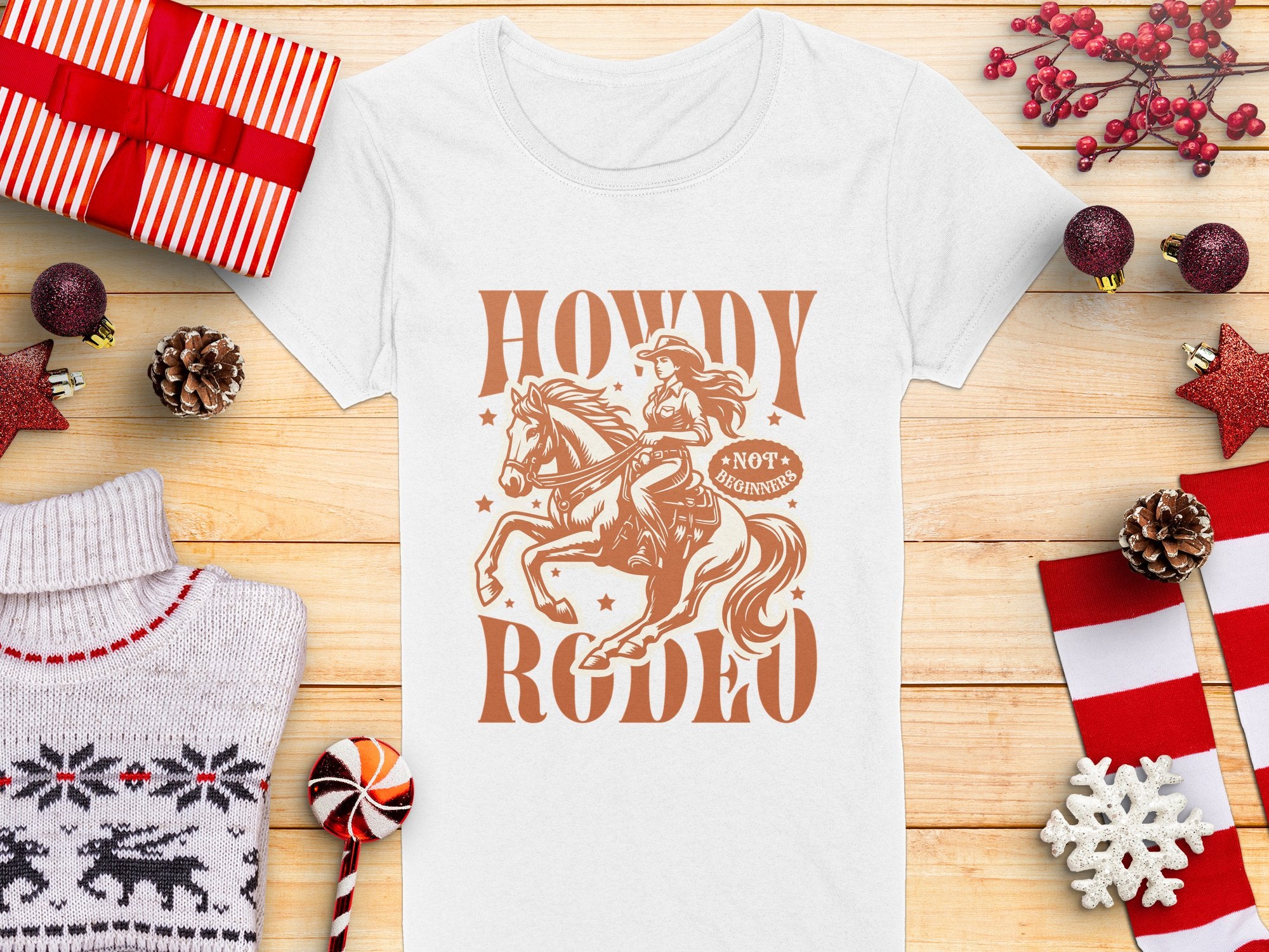 Womens white Garment Graphics T-shirt with a cowboy and horse, Howdy Rodeo text, and a Not Mounted tag. Styled with wrapped gifts, pinecones, sweater, candy cane, and holiday decor on wood.