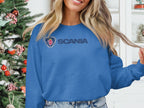 A person sporting a blue Garment Graphics Scania logo sweatshirt stands indoors with long hair and a slight smile. A decorated Christmas tree is in the background. Perfect for casual outings, this unisex sweatshirt ensures everyone can enjoy its comfort and style.