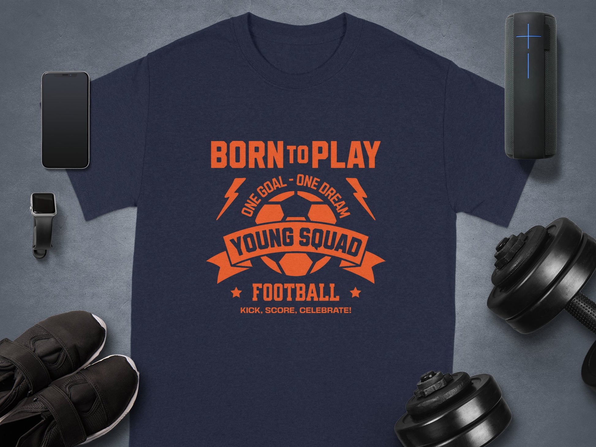 A dark blue Born to Play, One Goal - One Dream, Young Squad Football T-shirt by Garment Graphics features a vibrant orange graphic. It is artistically set among a smartphone, smartwatch, sneakers, dumbbells, and a portable speaker on a sleek gray background.