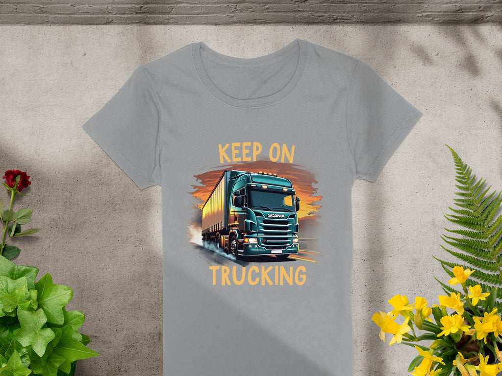 The Garment Graphics gray T-shirt showcases a bold truck graphic with Keep On Trucking, laid on concrete. Ideal for trucking fans, it features a red flower on the left and vibrant yellow flowers on the right.