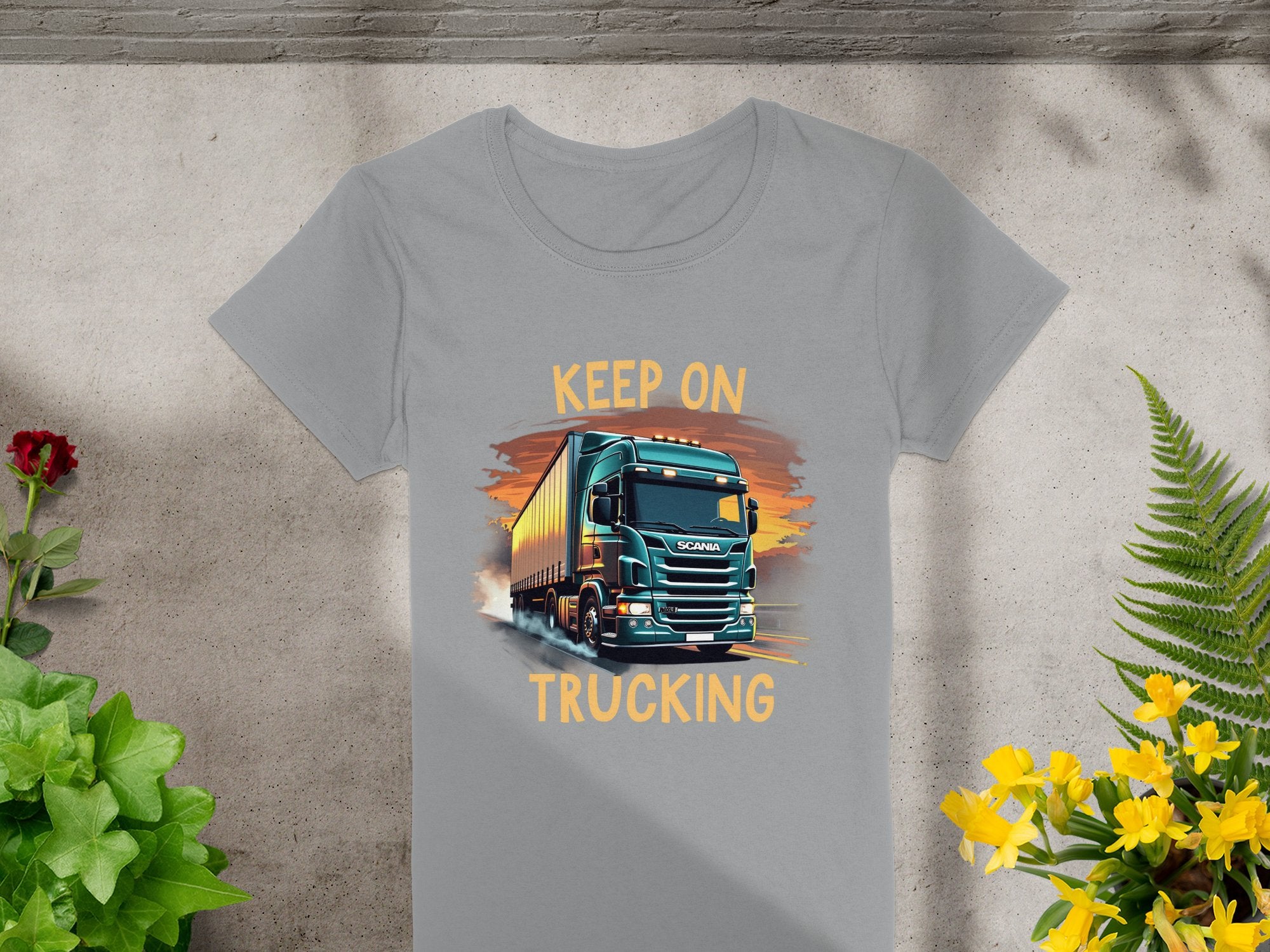 The Garment Graphics gray T-shirt showcases a bold truck graphic with Keep On Trucking, laid on concrete. Ideal for trucking fans, it features a red flower on the left and vibrant yellow flowers on the right.