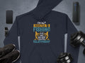 Flat lay of a dark fishing hoodie from Garment Graphics featuring the vibrant text Im not addicted to fishing, we are just in a very committed relationship in blue, white, and orange. The unique design is surrounded by essentials like a smartphone, smartwatch, sneakers, dumbbells, and a portable speaker.