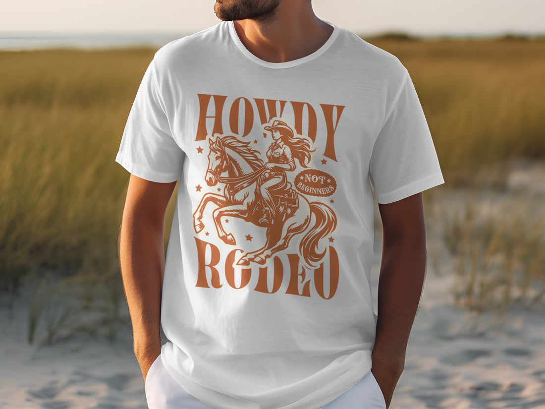 A person is wearing a white Garment Graphics T-Shirt featuring a brown cowboy on a horse with the text Howdy Rodeo and Not Branded. The background of grass and sand suggests a beach or dune, ideal for western culture fans.