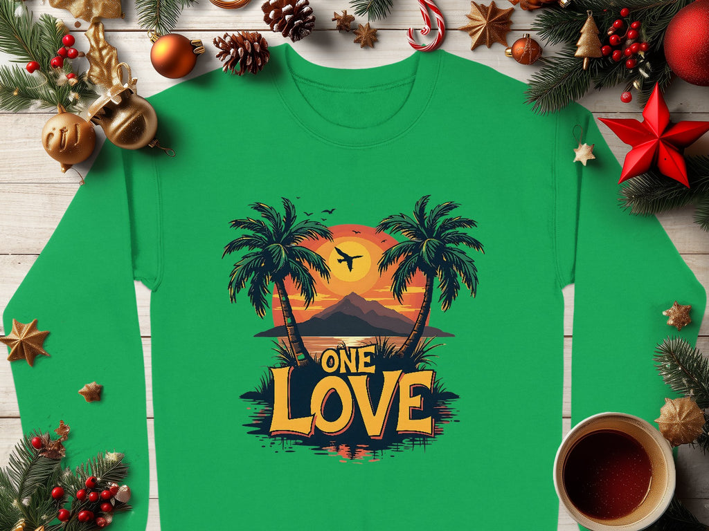 A Garment Graphics unisex green shirt, with a sunset between palm trees and One Love, sits on a wooden surface decorated with holiday ornaments, pinecones, baubles, and a steaming coffee.
