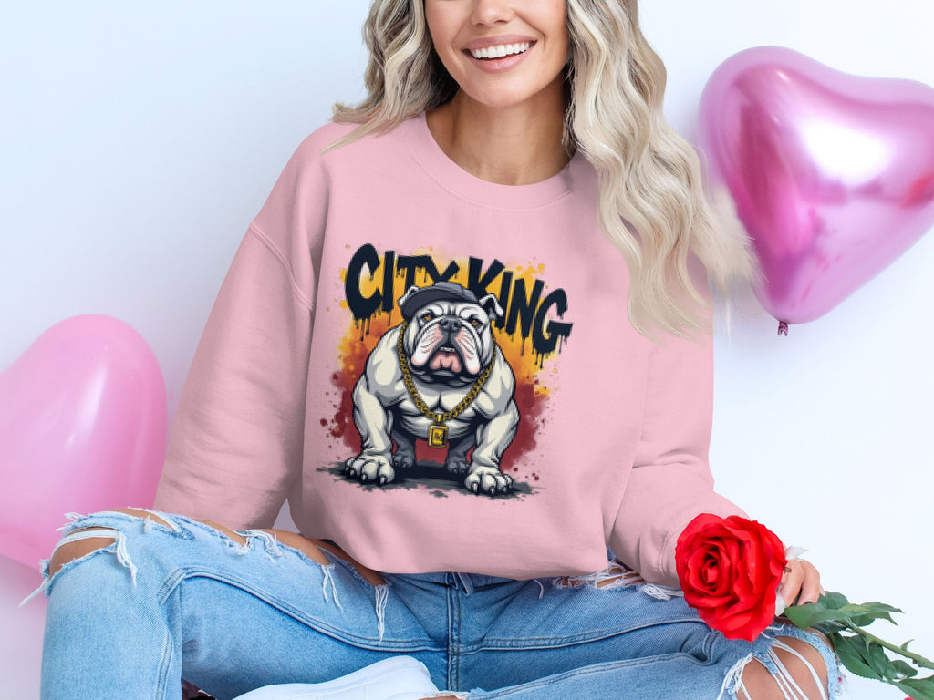 A person wearing a Garment Graphics pink City King sweatshirt with a bulldog graphic and gold chain sits cross-legged. They smile, holding a red rose, surrounded by two pink heart-shaped balloons, in their distressed jeans.