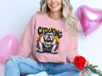 A person wearing a Garment Graphics pink City King sweatshirt with a bulldog graphic and gold chain sits cross-legged. They smile, holding a red rose, surrounded by two pink heart-shaped balloons, in their distressed jeans.