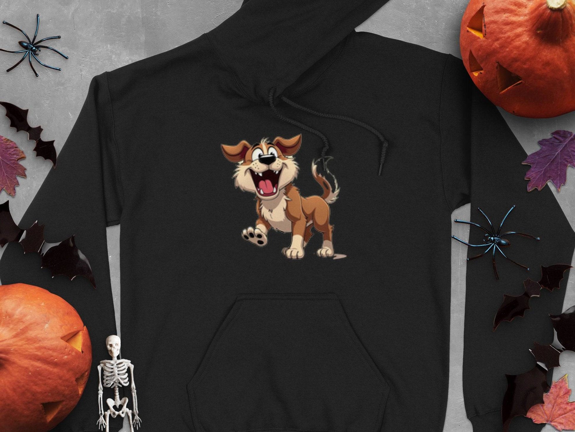 The Garment Graphics black cartoon puppy hoodie, ideal for dog lovers and a playful wardrobe piece, is surrounded by Halloween decor such as bats, spiders, pumpkins, and a small skeleton on a gray surface.