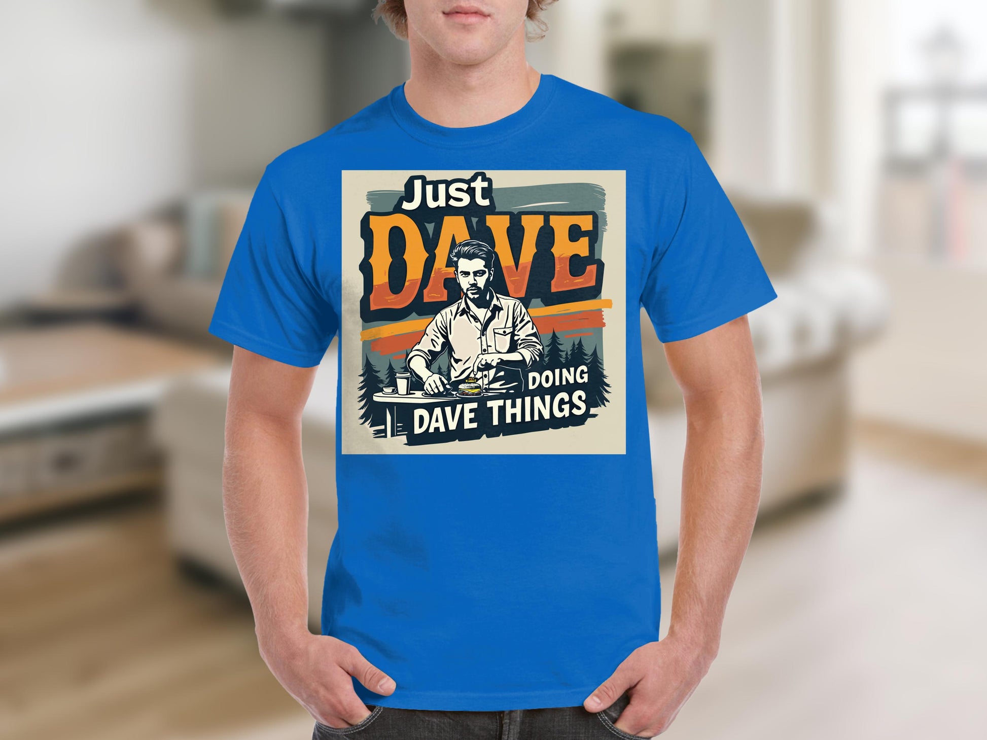 In a cozy indoor setting with furniture, a man models the blue graphic T-shirt by Garment Graphics. Its classic fit and features an illustration with the text Just Dave Doing Dave Things, highlighting his casual style.