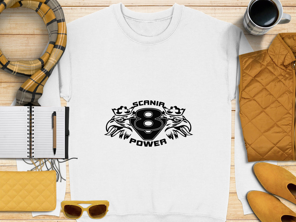 A Garment Graphics white sweatshirt with a black Scania Power V8 logo of two lions is displayed on wood, surrounded by a scarf, notebook, pen, sunglasses, yellow jacket, coffee cup, and purse.