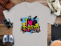 Garment Graphics presents a gray T-shirt with a vibrant graffiti-style heart and can icon, reading I Love Graffiti. Its perfectly staged on wood with a camera, binoculars, gloves, map, and leather pouch for street-art enthusiasts.