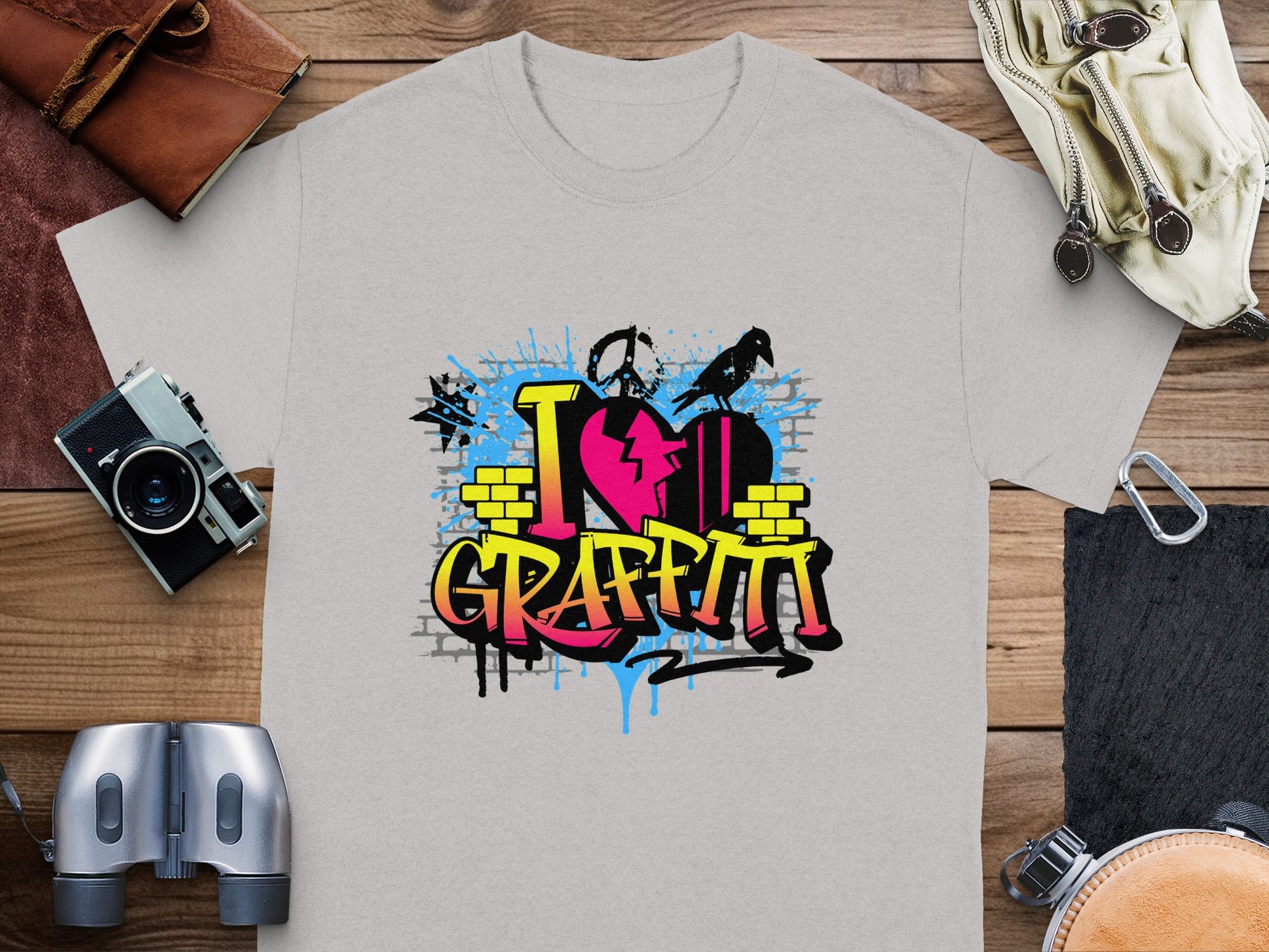 Garment Graphics presents a gray T-shirt with a vibrant graffiti-style heart and can icon, reading I Love Graffiti. Its perfectly staged on wood with a camera, binoculars, gloves, map, and leather pouch for street-art enthusiasts.