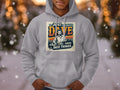 A person wears a Garment Graphics hoodie in medium-heavy fabric, with Just Dave Doing Dave Things boldly printed on gray. It features an illustration of someone in sunglasses amidst gently falling snowflakes.