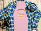 On a wooden surface lies a Garment Graphics pink Father and Son Fishing Partners for Life T-shirt, symbolizing family bonding, surrounded by a blue plaid shirt, headphones, a camera, and a coffee cup lid.