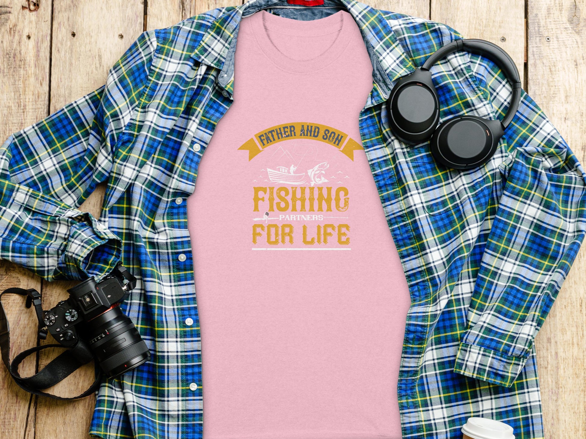 On a wooden surface lies a Garment Graphics pink Father and Son Fishing Partners for Life T-shirt, symbolizing family bonding, surrounded by a blue plaid shirt, headphones, a camera, and a coffee cup lid.