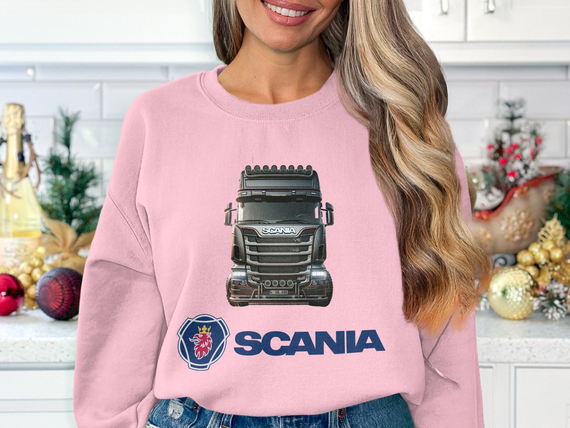 In a cozy kitchen, a truck enthusiast dons Garment Graphics vibrant pink crewneck sweatshirt, showcasing a Scania truck and logo surrounded by festive ornaments and charming decor.