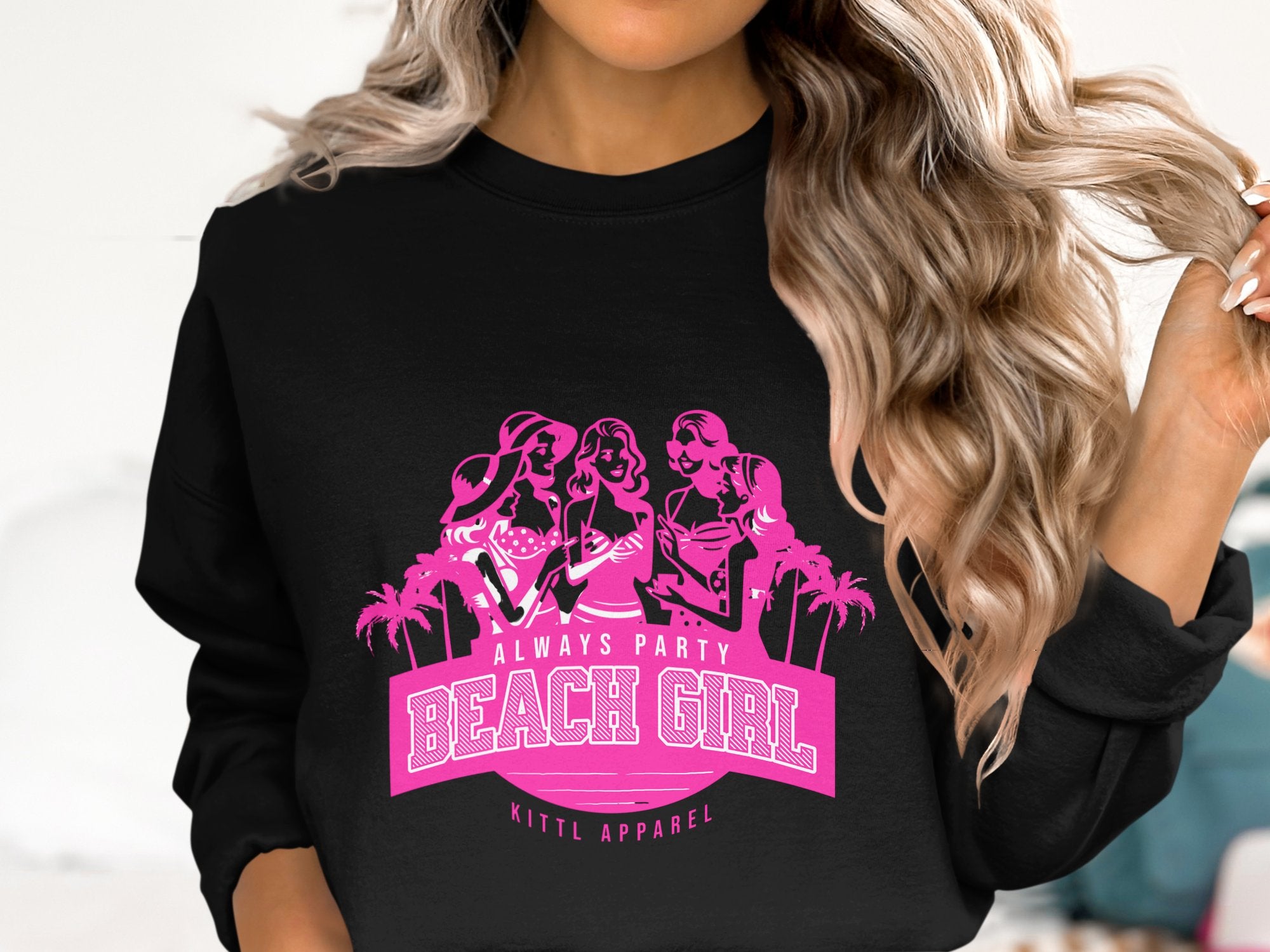 A person with wavy, long blonde hair is wearing a black sweatshirt from Garment Graphics featuring a pink graphic of stylized women, palm trees, and the text Always Party Beach Girl.