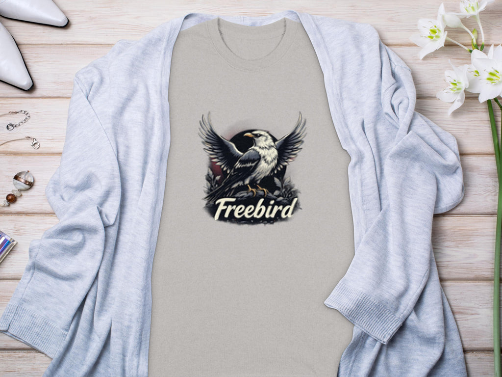 A beige 100% cotton t-shirt by Garment Graphics showcases a striking bird graphic with Freebird beneath. Styled with a light gray cardigan, flowers, and white shoes on a wooden surface, it enhances the Freebird eagle T-Shirt’s look.