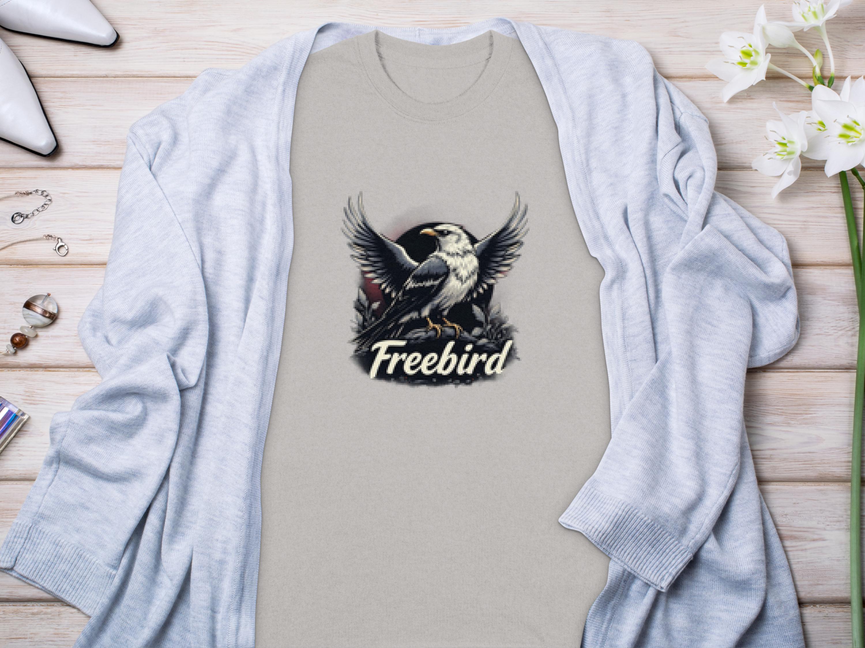 A beige 100% cotton t-shirt by Garment Graphics showcases a striking bird graphic with Freebird beneath. Styled with a light gray cardigan, flowers, and white shoes on a wooden surface, it enhances the Freebird eagle T-Shirt’s look.