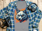 A gray sweatshirt from Garment Graphics, featuring a fierce wolf graphic with orange and blue accents, lies on wood. It exudes a wild side alongside a blue plaid shirt, headphones, a camera, and a white coffee cup.